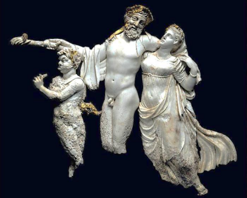 Das Bild zeigt eine hellenistische Statue auf schwarzem Hintergrund. Darauf sind drei Figuren zu sehen. Ein Satyr, ein Mann und eine Frau.