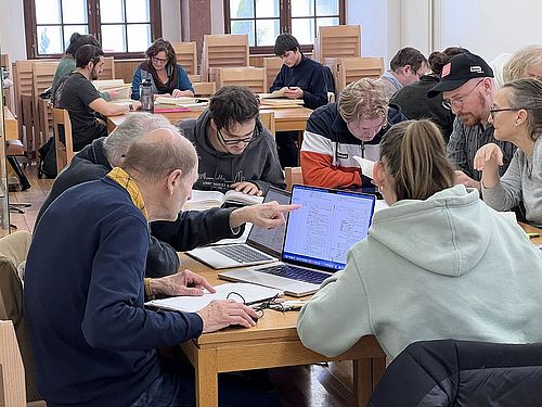 Das Bild zeigt mehrere Studierende, welche nahe aufeinander sitzen und gemeinsam, mithilfe von Nachschlagewerken, akkadische Texte übersetzen.