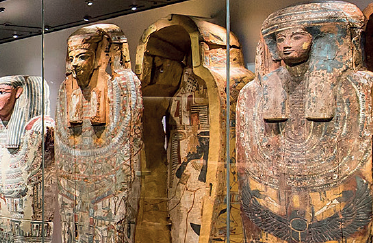 Das Bild zeigt an eine Reihe an Sarkophage, welche hinter einer Vitrine stehen.